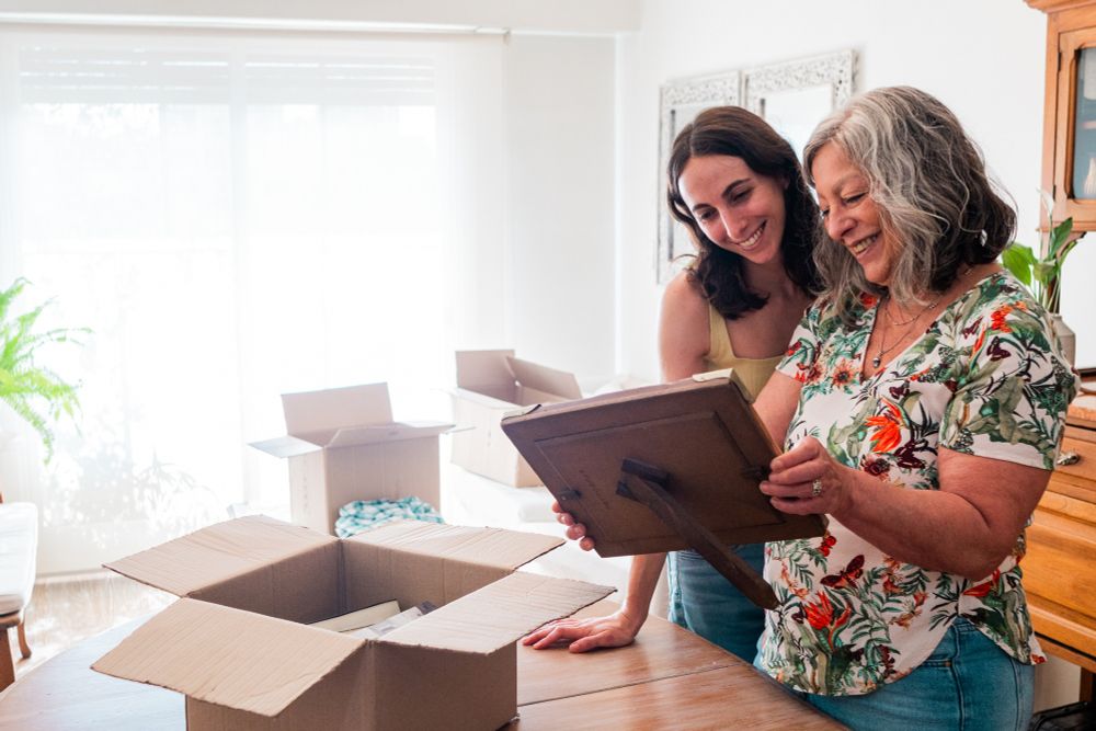 Two women unpacking boxes in a bright room, one holding a framed picture while the other looks on.
