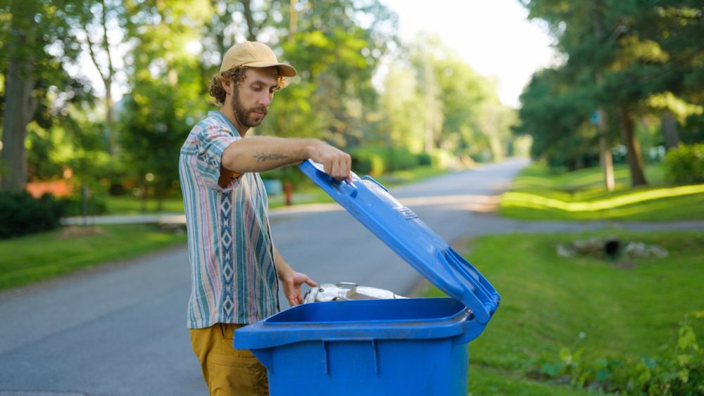 Man placing a clear plastic bag of recycle into a blue bin.