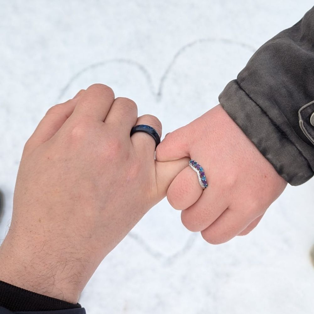 Rain and their partner Ape wearing their engagement rings. Ape's ring is a black band with blue details. Rain's ring is a silver band with heartshaped gems alternating blue and purple. In the background is a heart drawn in the snow.