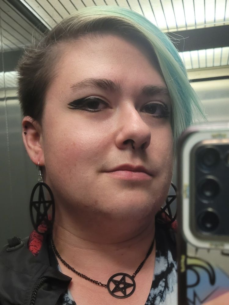 Selfie of Rain with black winged eyeliner, black pentagram earrings and necklace, and black septum and helix piercings. Their hair is faded to greenish blue and the side of their head is shaved but growing out.