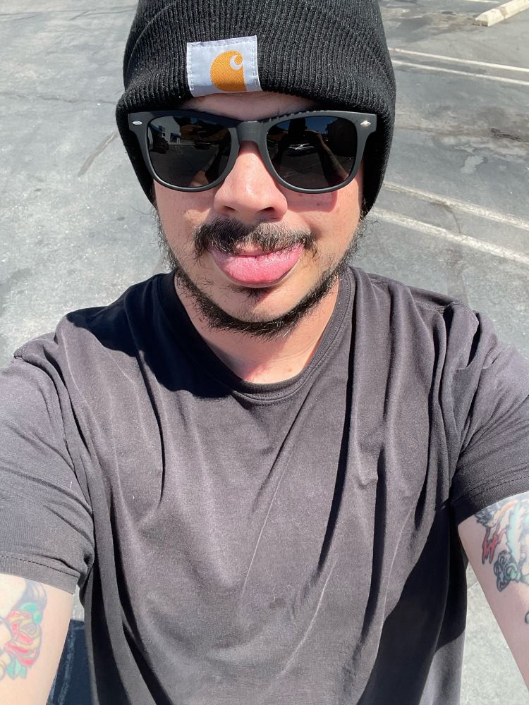 Selfie of Zack in a sunny outdoor parking lot. His tongue is sticking out and he is wearing sunglasses and a beanie, dressed in black