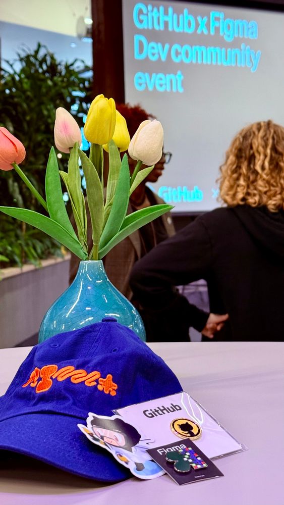 On a table with a vase with tulips are some swag placed: Blue hat with orange embroidery print, github pin in black and yellow, sciencey github sticker and figma pin in multiple colors on one side and shiny on the other.