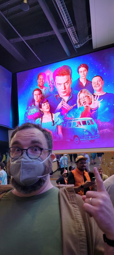 Dom in front of the Dimension 20 promo art.