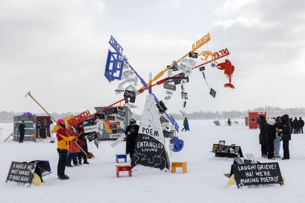 A whimsical structure sits on the ice with cloudy skies behind. On the structure is written "A Poem for Entangled Living," and many words are written on colorful posts that blow in the wind. 
