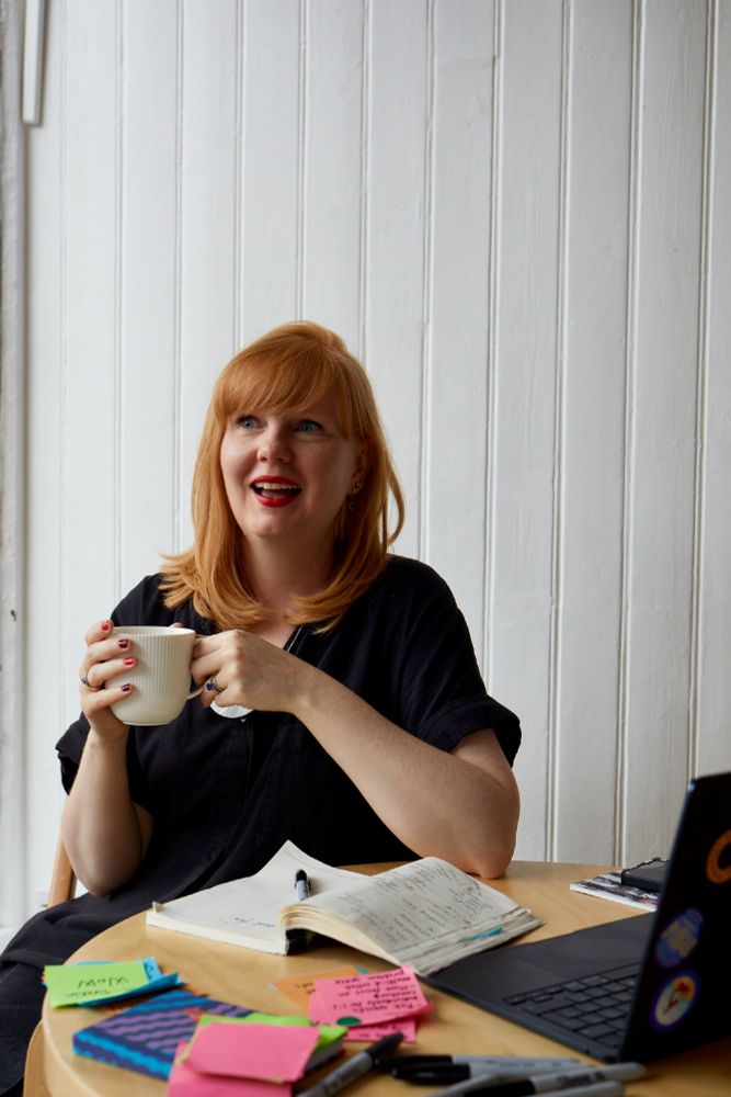 Woman with ginger hair sips a coffee smiling with surrounded by notebooks, post-its notes and a laptop covered in stickers