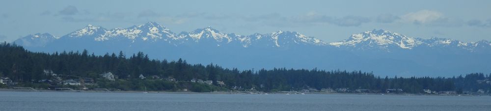 Rocky, snow capped peaks line the distant horizon. In front of those, much closer, is a line of trees, with a line of houses along a shore. The bottom 1/5 of the photo is water, between the shoreline and the camera.