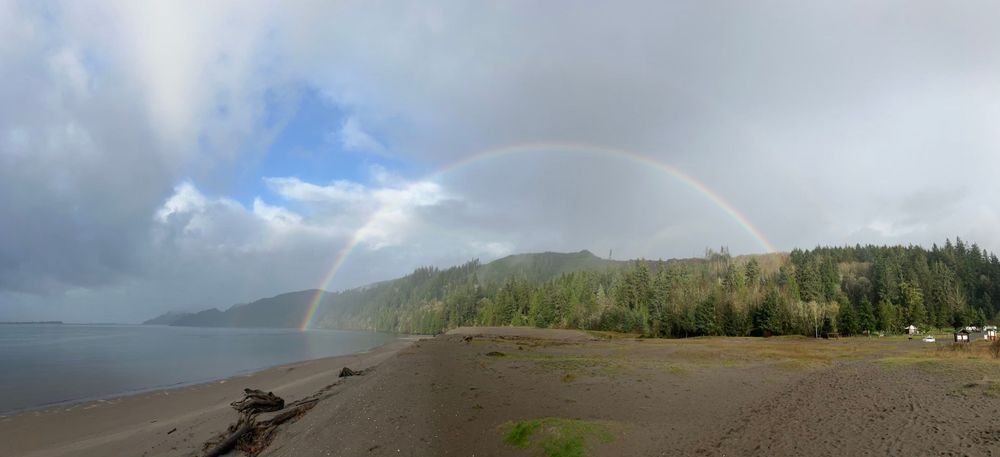 A large body of water, without an opposite shore visible, takes up the left 1/3 of the photo. A forested ridge extends from left to right, behind the water and all the way across to the right side. There is a beach in the foreground that takes up the front 1/3 of the photo, from the left 2/3 to the right edge. The top 1/3 of the photo is mostly grey sky, but a small hole of blue sky is towards the left 1/3 of the photo. A rainbow extends from the left 1/3 to the right 1/3 of the sky, in a near 180 degree arch.