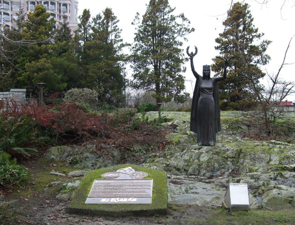 A monument with a sculpture of a woman holding a wreath and eagle is on a small basalt outcropping. An interpretive sign is in the foreground. The area around it is partially forested, though there is a very large building in the background.
