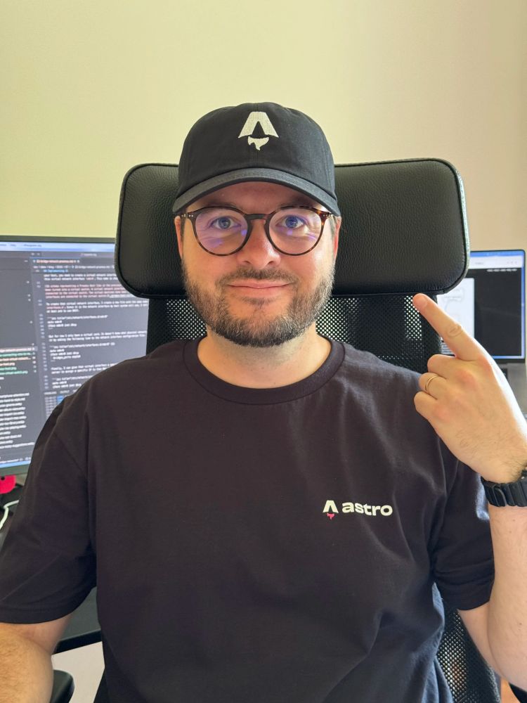 A picture of me, a while man in his 30s, wearing a black cap and a black t-shirt with the Astro logo. I'm also wearing glasses. I'm smiling and my fingers points at my face.

In the background there is a MacBook Pro and a stand and a large monitor with a code editor open.
