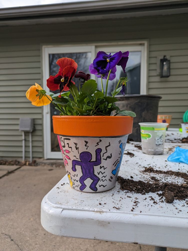 Small terracotta pot with colorful pansies inside. It is painted silver with a purple human figure in the cartoon style of Keith Haring 