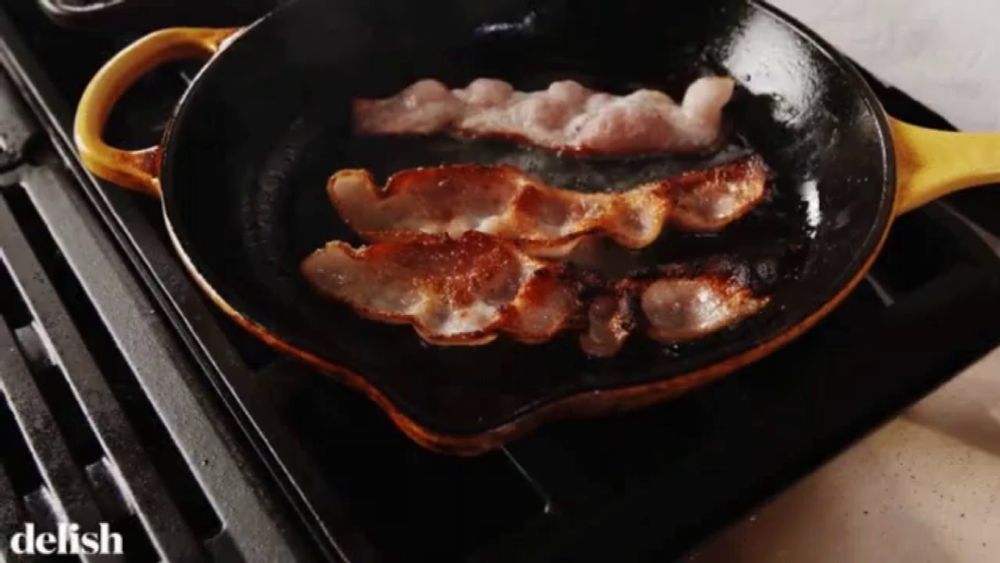 bacon is being cooked in a skillet on a stove with the word delish visible