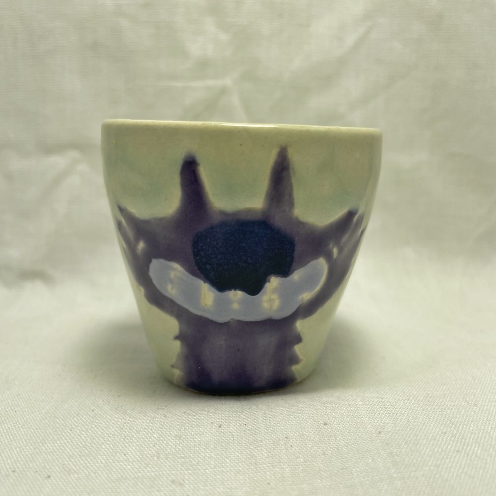 the same light green shot glass shown from the other side. it is decorated with a single, stylized purple wolf head, with a big toothy lavender grin and a big dark blue nose. the glaze has run together in a couple areas. 