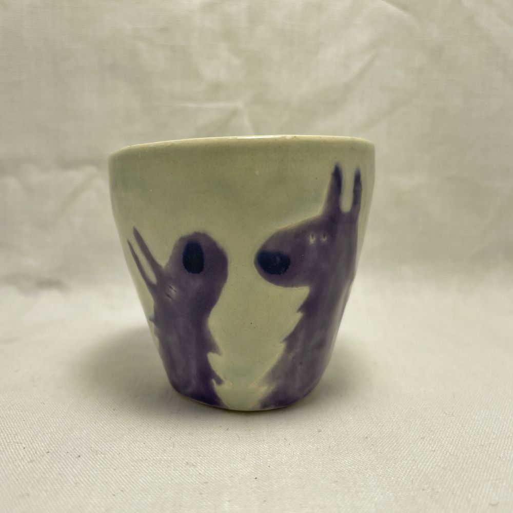 a light green wheel thrown shot glass decorated with two stylized purple wolves, shown in portrait looking at each other. they both have big, dark blue noses.