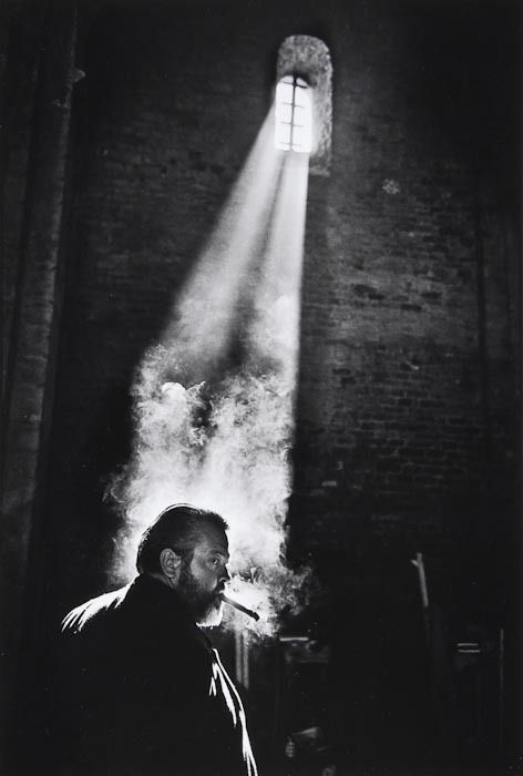 portrait B&W photo of Orson Welles in profile smoking a cigar in the lower left third of the frame, spotlit by light coming through a small window set into a brick wall high above him