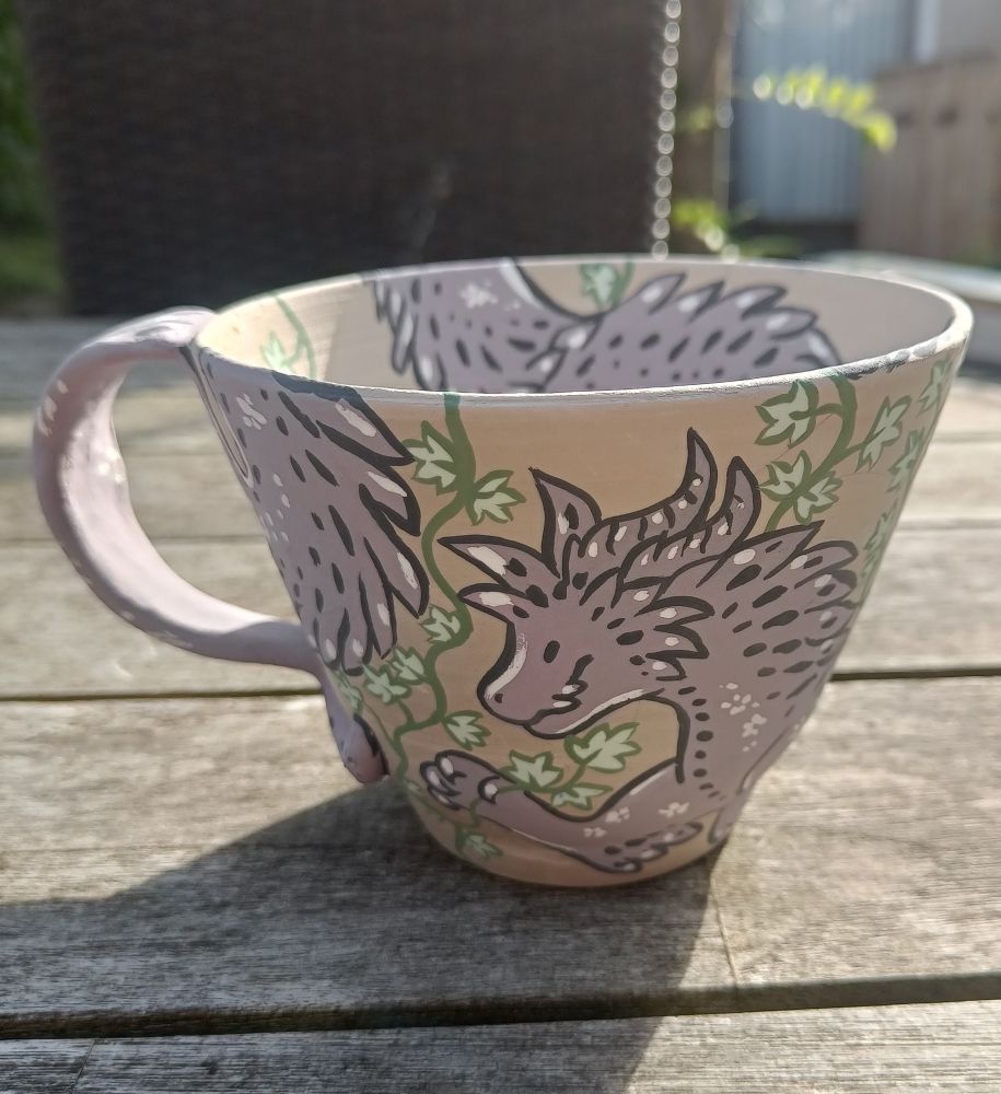 A ceramic mug, unfired, sits atop a wooden table. It features a spiky, greyish-purple dragon holding a string of leafs. It's design is inspired by a bramble.