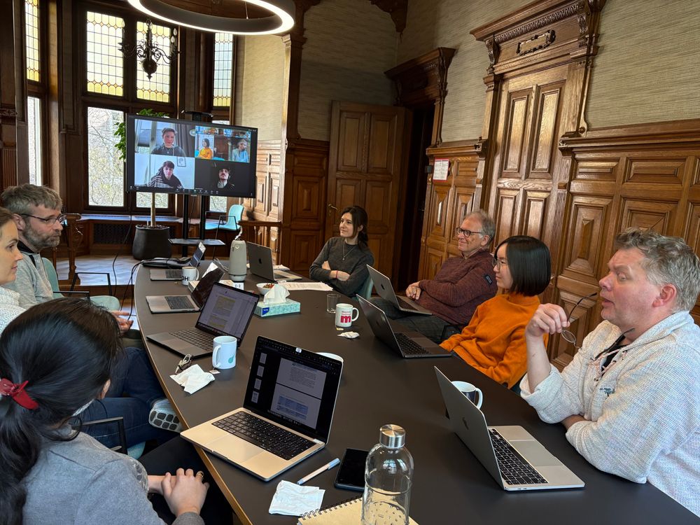 People sitting behind laptops around a table in a meeting room with wood paneling and several people who are attending the meeting remotely via a screen 