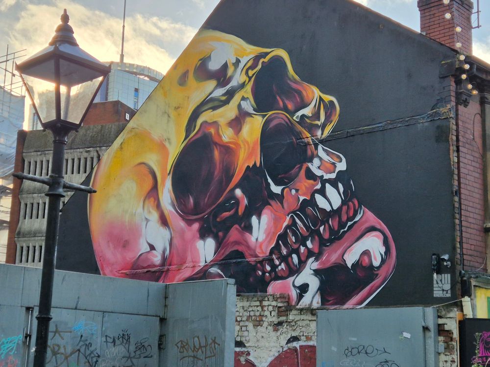 A street painting on the side of a building of a human skull. It is yellow and pink in colour on a black background