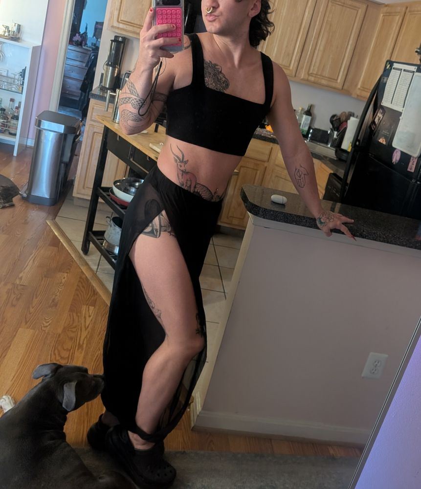 Hedonix stands at 3/4 wearing a black crop tank top and showing off their muscular leg peeking out of sheer harem pants. 