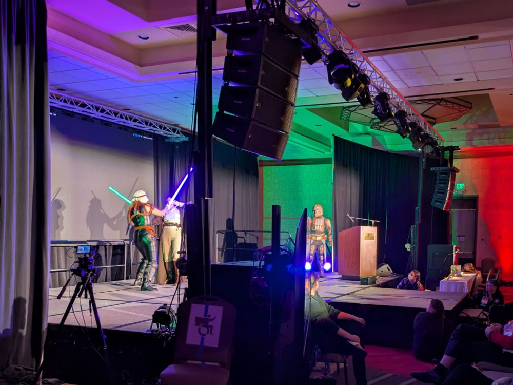 A photo of two performers having a light saber battle on stage. A green light glints off of one performer's shiny outfit, matching her light saber.