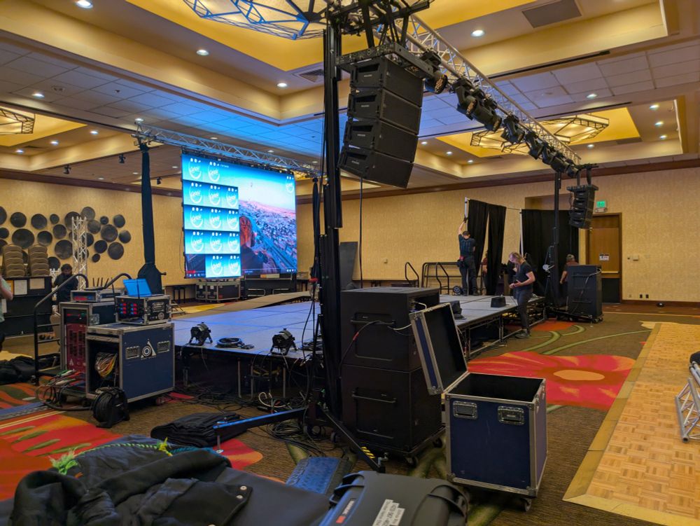 A photograph of main stage while it was being set up. There is a downstage truss with lights and speakers, and an upstage truss with an LED wall being configured. People in the background are setting up drape.