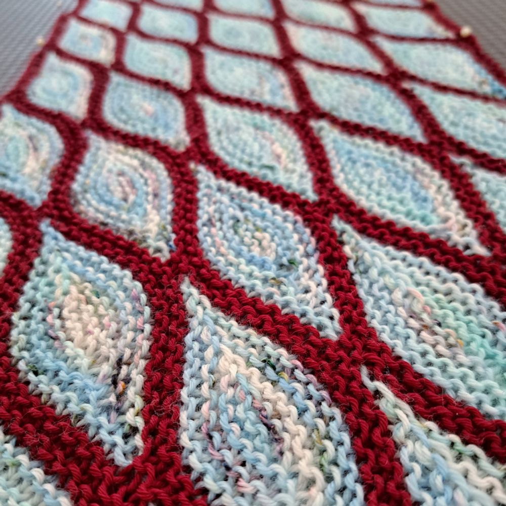 Hand-knitted cowl knitted on the bias pinned on a blocking board. The cowl is made up of very light blue garter stitch short row diamond within dark red contour rows.