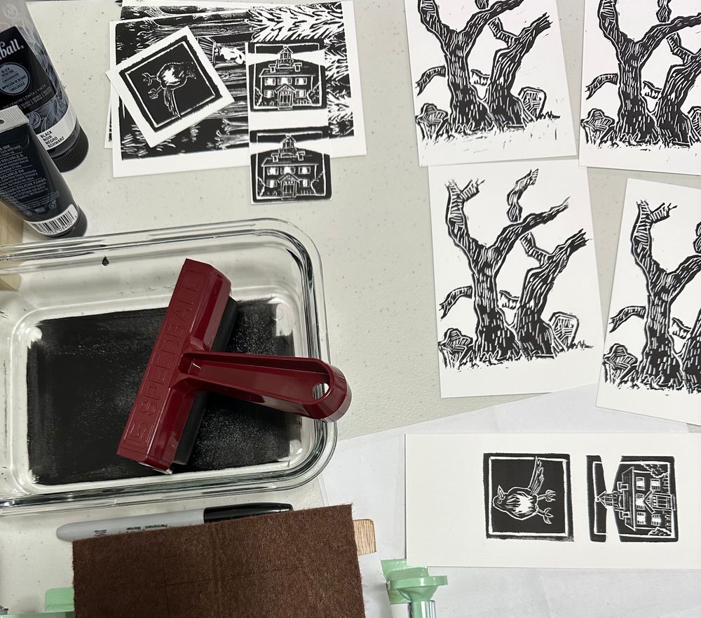 View of a workspace from above. A small print press partially in view lies open, and a brayer rests in a glass pan of ink. Scattered around these tools are ink bottles, scattered papers, and prints depicting magpies, lighthouses, forests, and trees with gravestones near their base.