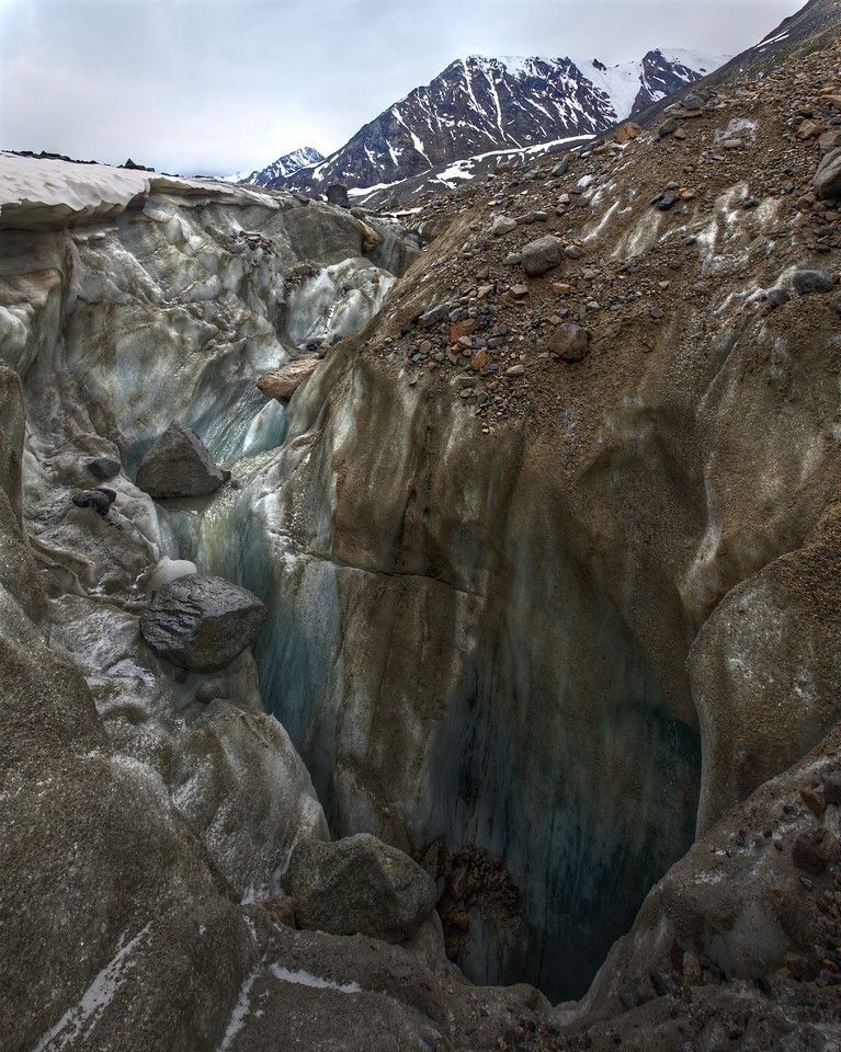 A deep, wide moulin carved into the ice of the Black Rapids Glacier. The walls are a mix of dirty gray and clean blue ice, streaked with meltwater channels and scattered rocks. Large boulders sit on the ice near the opening, and the moulin drops steeply out of sight. In the background, snow-covered peaks of the Eastern Alaska Range rise under an overcast sky.