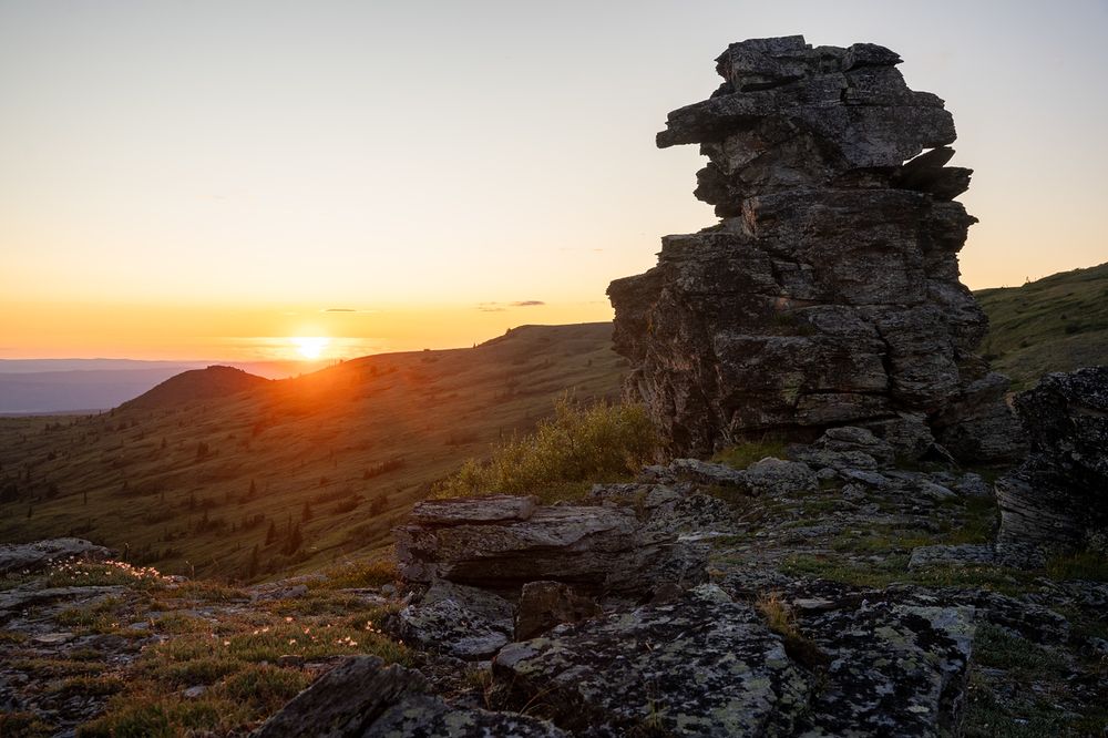 Sunset over the rolling tundra hills of Wickersham Dome in interior Alaska. The sun is low on the horizon, casting golden-orange light across the landscape. In the foreground, a large, weathered rock formation stands tall on the right, its blocky surface catching soft highlights from the setting sun. The terrain slopes gently into the distance, dotted with small spruce trees and patches of late-summer vegetation. The sky is mostly clear with a warm gradient from orange near the sun to pale blue higher up.