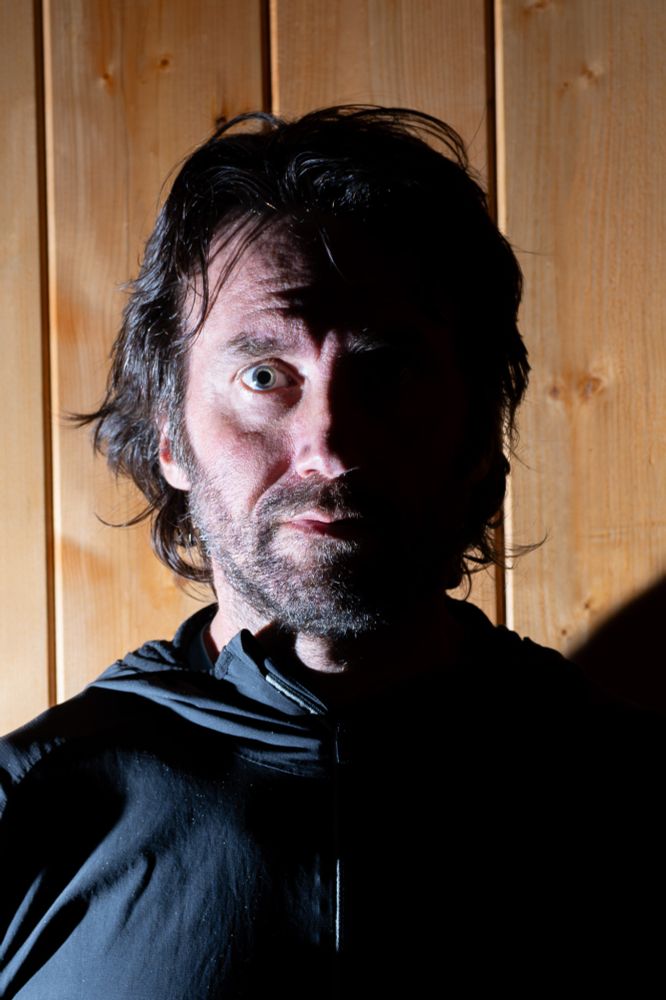 A man with medium-length dark hair and a short beard stands against a vertical wooden plank wall. His face is lit starkly from the right side, leaving the left side in deep shadow, with only one eye brightly illuminated. He wears a dark zip-up hoodie, and his expression is wide-eyed and intense, as if caught mid-thought or startled. The dramatic side lighting creates strong contrast and sharp edges in the shadows.