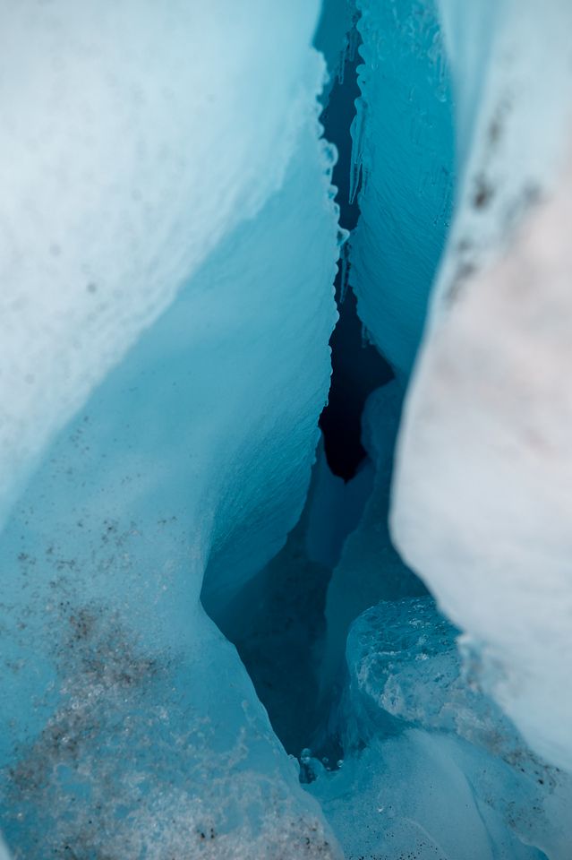 A close-up view inside a moulin on the Gulkana Glacier in the Eastern Alaska Range. The narrow opening reveals smooth walls of deep blue ice, fading into darker shadows as the moulin descends out of sight. The ice surface shows subtle textures and curves, with small bubbles and pockets visible, giving a sense of depth and coldness.