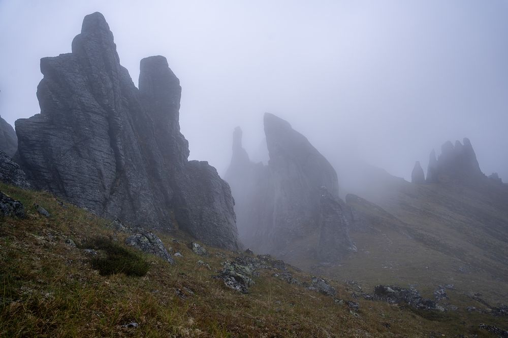 A foggy alpine scene on the descent from Mt. Prindle in interior Alaska. Jagged granite tors—tall, weathered rock spires—rise dramatically from the sloped tundra, partially obscured by thick clouds. The gray stone surfaces appear cold and damp, slick with moisture from the fog. The landscape slopes downward from left to right, covered with patches of alpine grasses, mosses, and scattered lichen-covered boulders. The massive stone towers fade into the distance, becoming silhouettes in the mist. The atmosphere is quiet and surreal, with low visibility and diffused light giving the whole scene a soft, eerie feel.