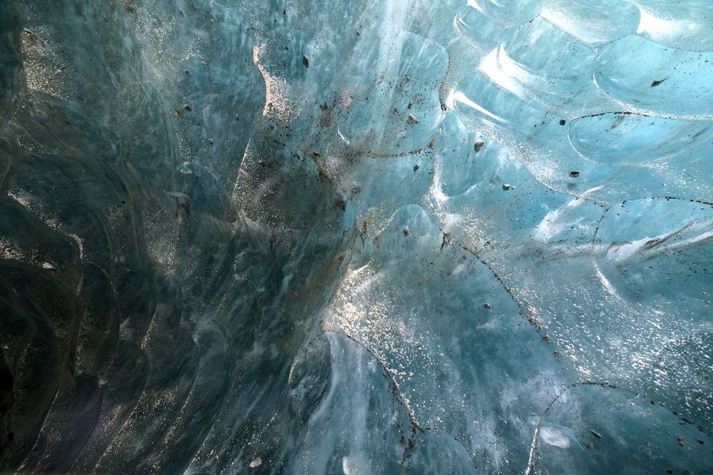Close-up view of the interior wall of a glacier cave, showing smooth, curved blue ice with streaks of trapped sediment and scattered small rocks embedded in the surface. Flecks of white frost and fine air bubbles create a speckled texture across the ice. Light falls unevenly across the wall, leaving the left side darker and the right side brighter, highlighting the sculpted, wave-like forms.