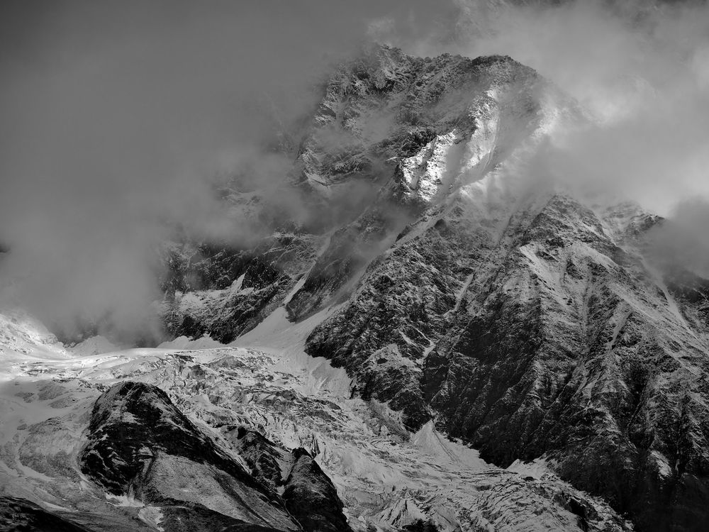 A black and white photo of an alpine icefall and steep, snow-dusted rock faces partially obscured by fog. A narrow shaft of sunlight breaks through the clouds, highlighting a section of the mountain slope and the fractured ice below. The terrain is sharply textured, with jagged ridgelines and deep crevasses visible where a tributary glacier meets the main Black Rapids Glacier.