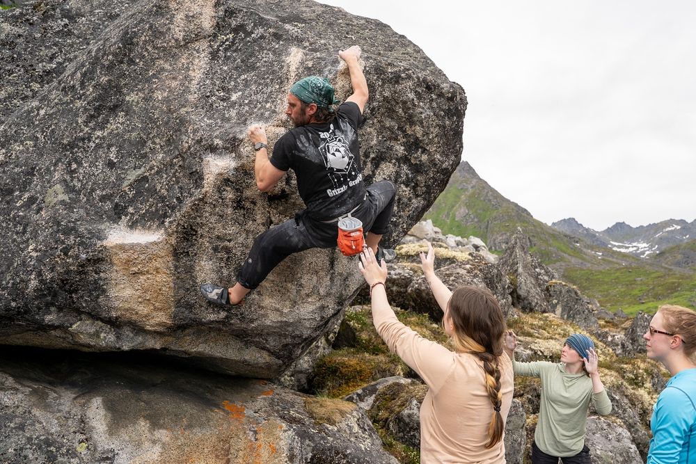 Man clings to the side of a large boulder mid-climb, feet smeared on small footholds, left hand gripping a chalked edge. Three climbers below spot him, arms raised for safety.