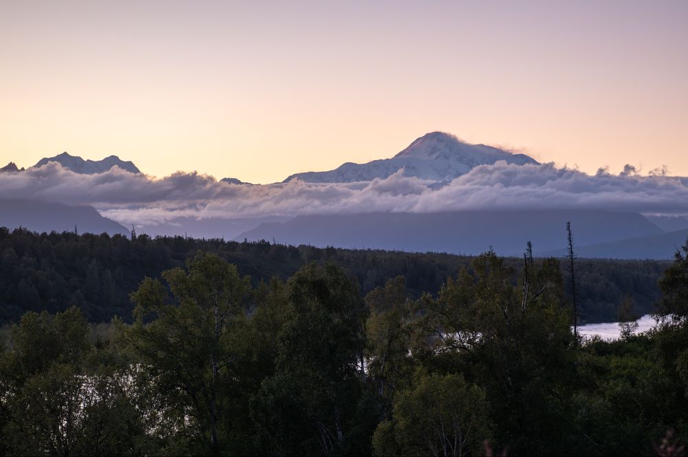 A sweeping view of Mount Denali at dusk, partially obscured by a band of mid-elevation clouds. The sun has set, leaving a gradient sky that shifts from soft peach near the horizon to pale lavender higher above. Below the mountain, a dark forested ridgeline and the reflective surface of a river add depth to the scene.