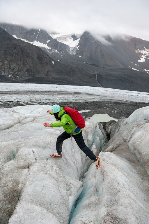 A person in a bright green jacket, red backpack, and teal hat steps across a narrow crevasse in glacier ice. The ice is gray-white with cracks running through it, and the background shows a wide glacier stretching toward dark, cloud-covered mountains.