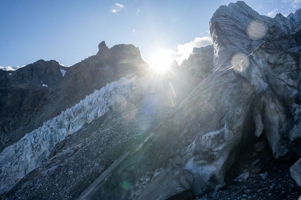 Sunlight shining over jagged mountain peaks and the Gabriel Icefall on the Gulkana Glacier in Alaska’s eastern Alaska Range. The icefall shows deep blue and white crevasses, with rocky debris scattered along its base. A towering formation of layered, weathered ice dominates the right foreground, while lens flare from the sun creates bright streaks across the image.