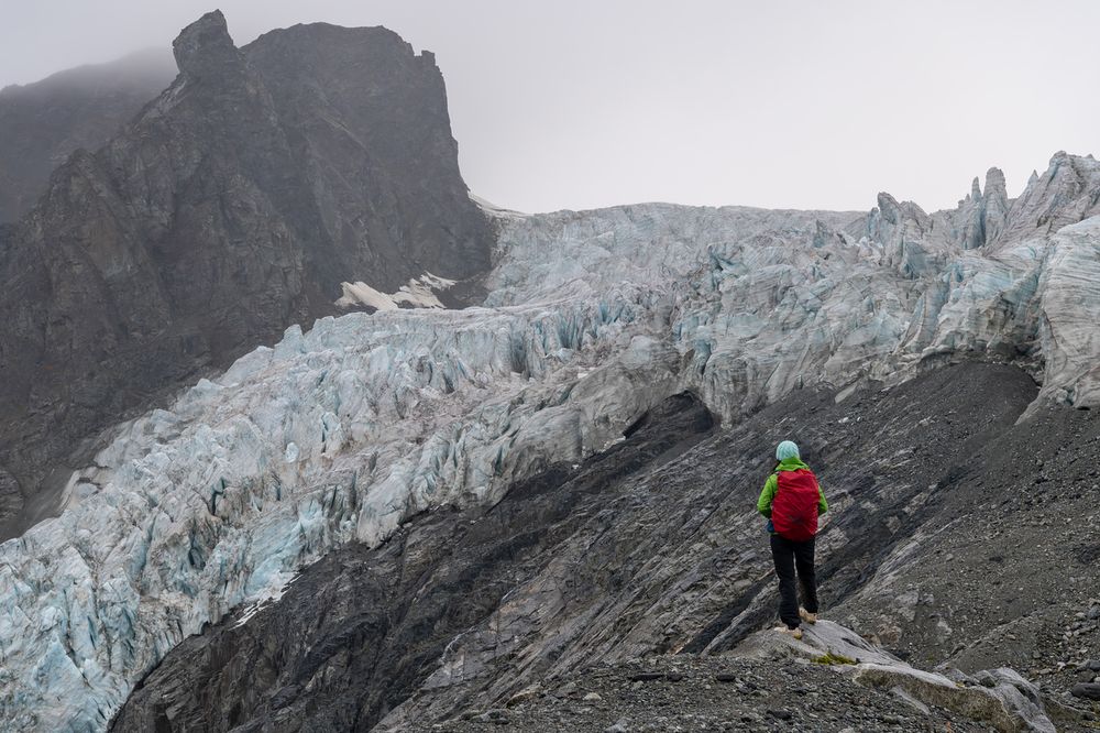 A person in a green jacket with a red backpack stands on rocky terrain, looking toward the fractured surface of the Gabriel Icefall on the Gulkana Glacier. The glacier ice is streaked with blue and white, pressed against a towering dark rock face. Mist and clouds obscure the upper slopes, adding to the stark, dramatic scene.