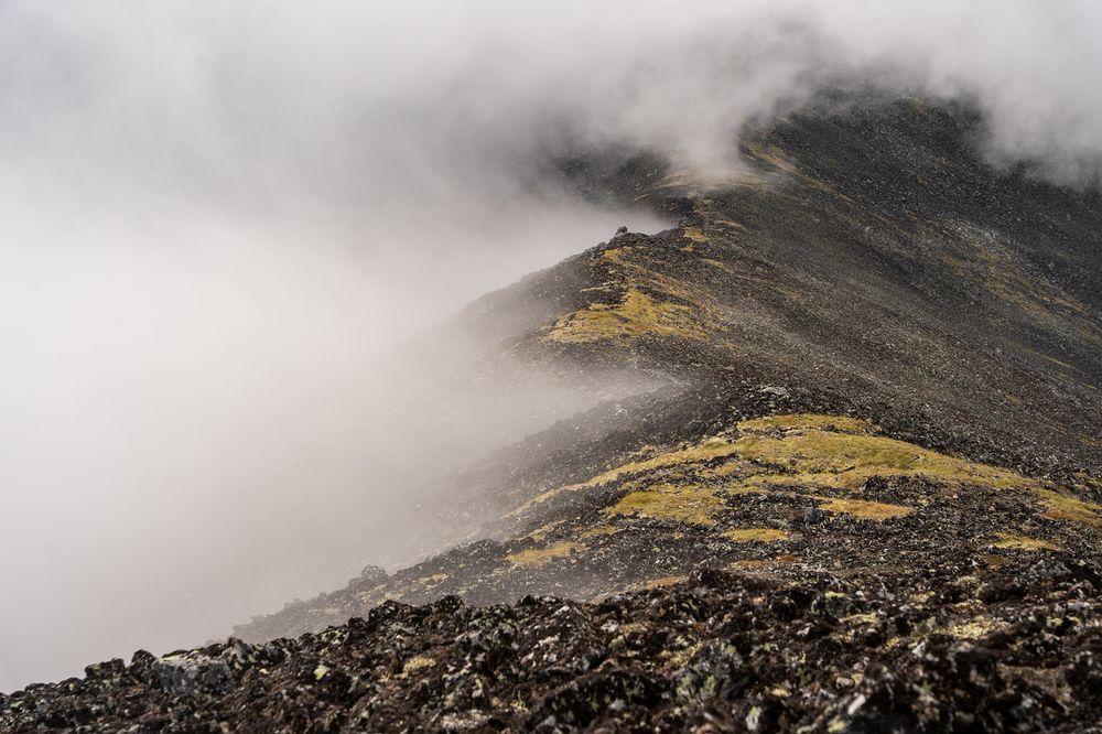 View from the summit of Mt. Prindle showing a dark, rocky ridge partially covered with patches of yellow-green tundra vegetation. Low clouds and mist sweep over the ridge, with visible condensation forming on the leeward side. The fog obscures much of the background, creating a soft, atmospheric effect against the sharp texture of the rocks in the foreground.