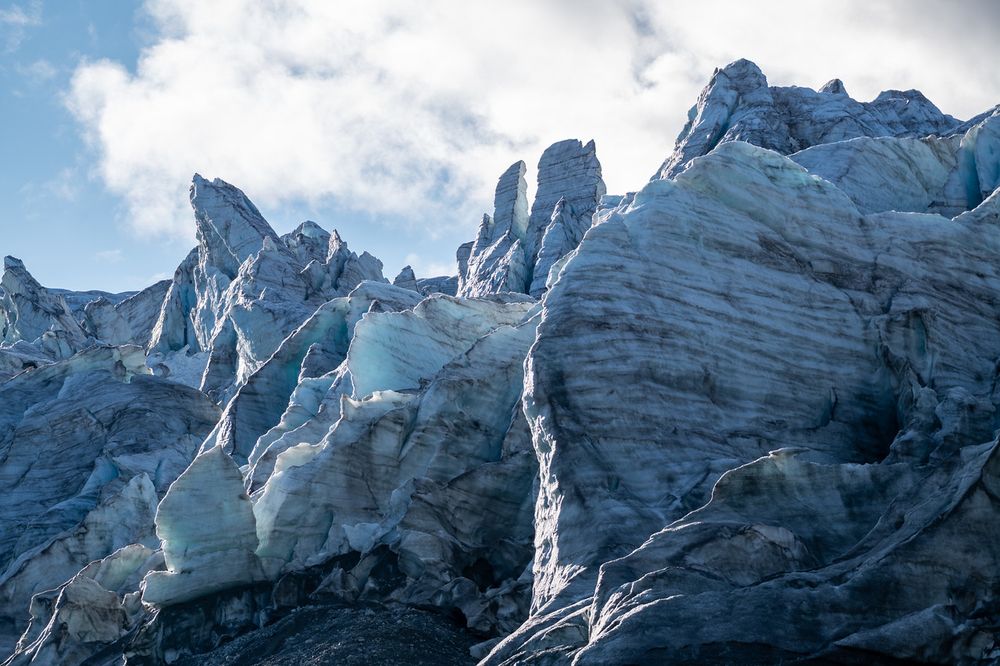 Dramatic ice formations in a glacier icefall, showing tall white and blue ice seracs and spires rising against a cloudy sky. The heavily crevassed ice displays complex fracture patterns and deep blue crevasses between the towering ice blocks.