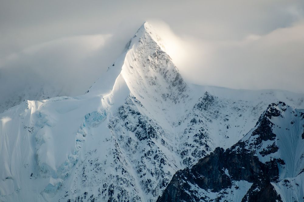 Snow-covered peak in the Alaska Range illuminated by low evening light, with jagged rock faces and glacial ice visible beneath the snow. Thin clouds partially obscure the summit.