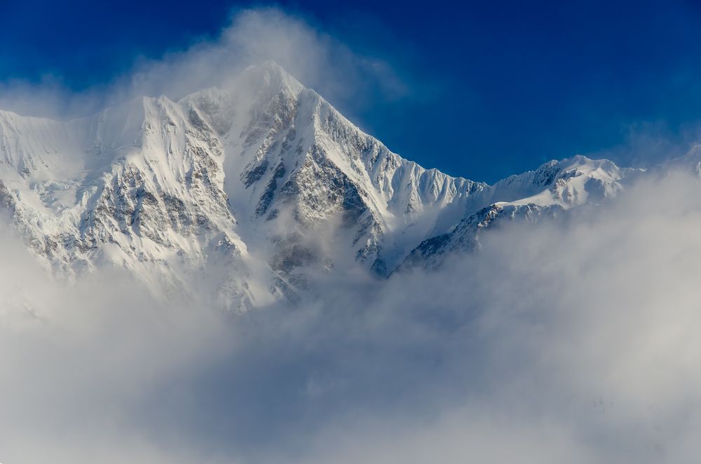 Snow-covered peak of McGinnis in the Alaska Range rises sharply behind drifting clouds. Wind lifts snow from the summit and ridgelines, creating a fine plume against a deep blue sky. The lower portion of the frame is filled with soft clouds partially obscuring the base of the mountain.
