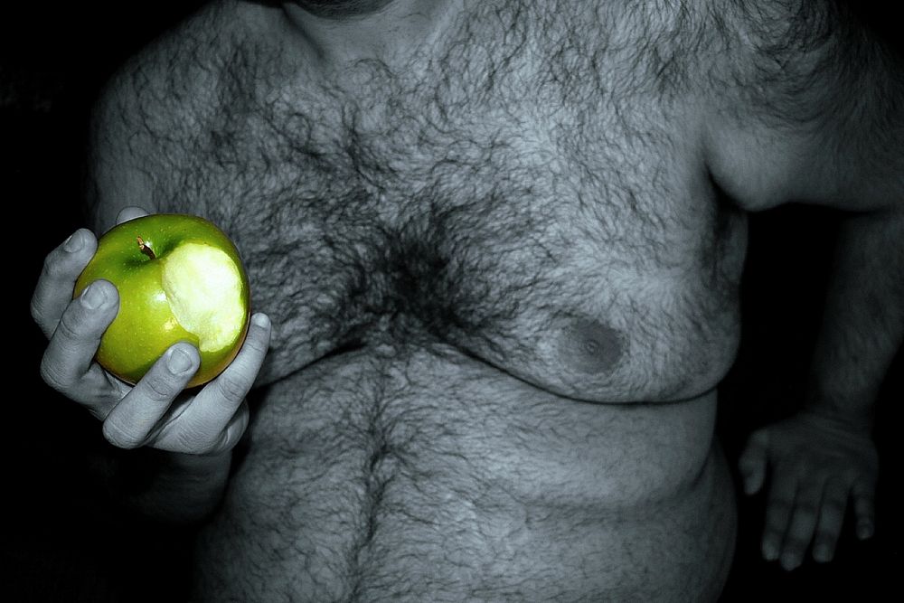 A black and white picture of a shirtless, hairy, fat man eating a granny Smith apple, which is in color.