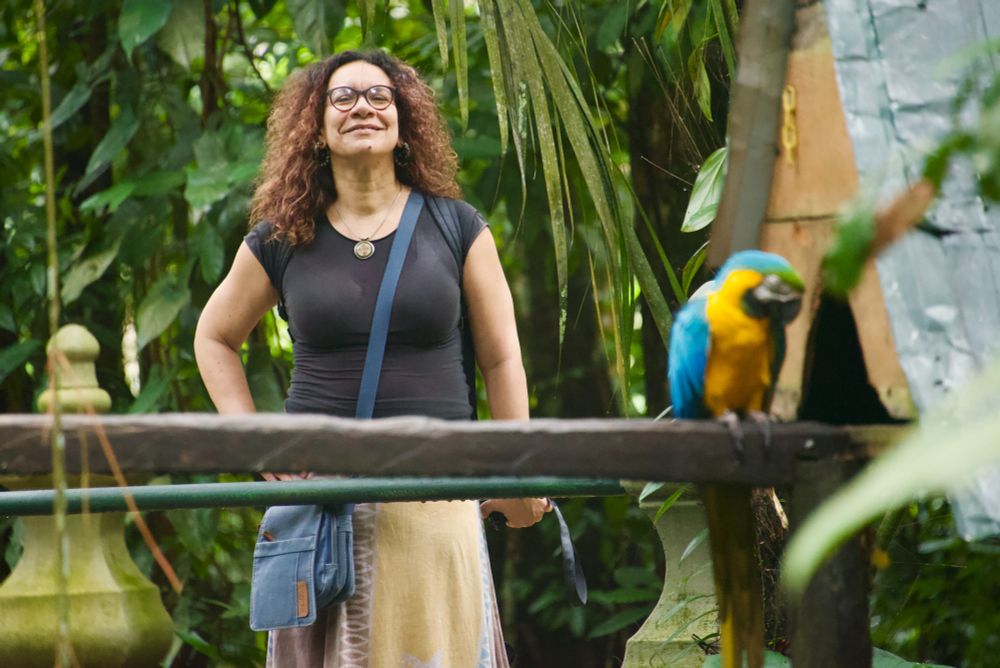 Woman of mixed Black heritage wearing glasses, black T-shirt & yellow skirt, with red dyed hair, in a park with a big parrot in the foreground. The macaw lives freely in the park & is very cheeky. 