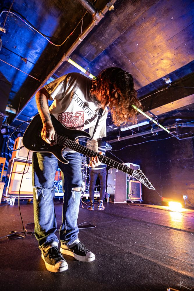 Upward angled photo of a guitarist bending at the hip, their long hair falling in their face. Warm tones with some purple and blues. 