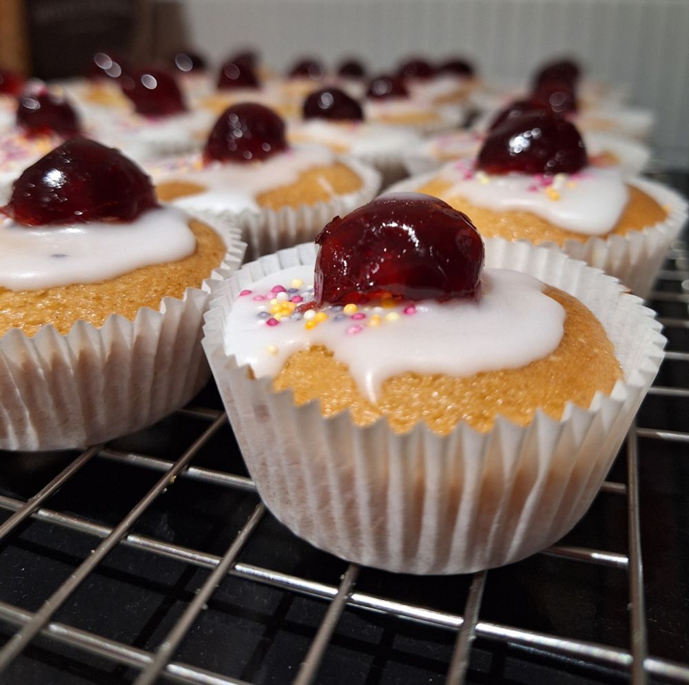 Fairy cakes in paper cases, decorated with white icing and cherries, on a wire rack.