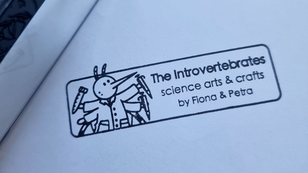photo of a black stamp on white paper, it shows a mosquito in a labcoat holding pipettes and a tube, and the text says "The Introvertebrates science arts & crafts by Fiona & Petra".