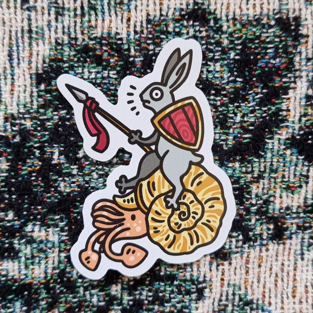 sticker on a pixelated looking blanket. the sticker has a medieval bunny holding a spear and red shield riding a yellow ammonite.