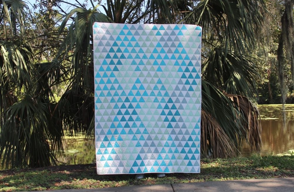 A large quilt in a pattern of diamond shapes against a white background. The diamonds are formed by 60 degree triangles in a various of blue, aqua, and teal solids alternating with white triangles. The quilt is being held up by an unseen person standing behind it. In the background are palm trees and a bayou. 