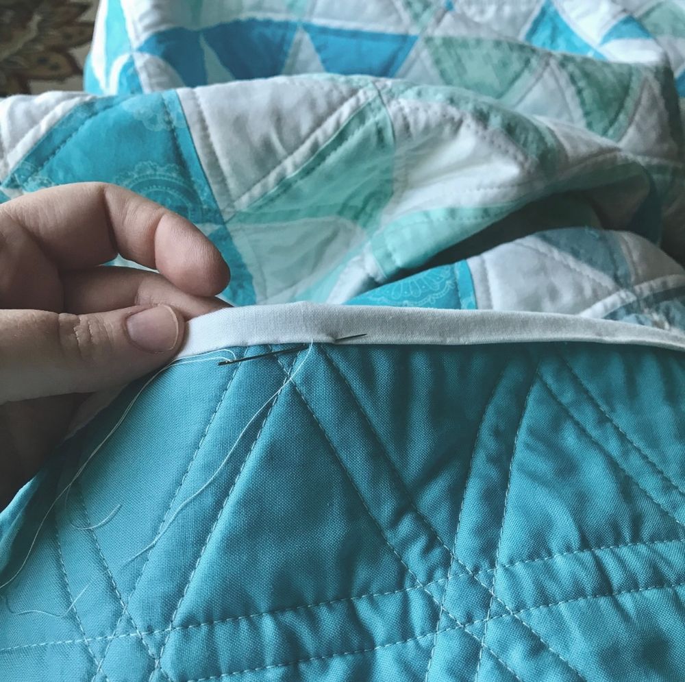 Photo of a crumpled blue and white quilt with one side flipped up to show the binding and back. White stitching on the solid blue back makes an interlocking triangle pattern. A hand holds the partially stitched white folded binding, pierced by a threaded needle where the stitching progresses. 